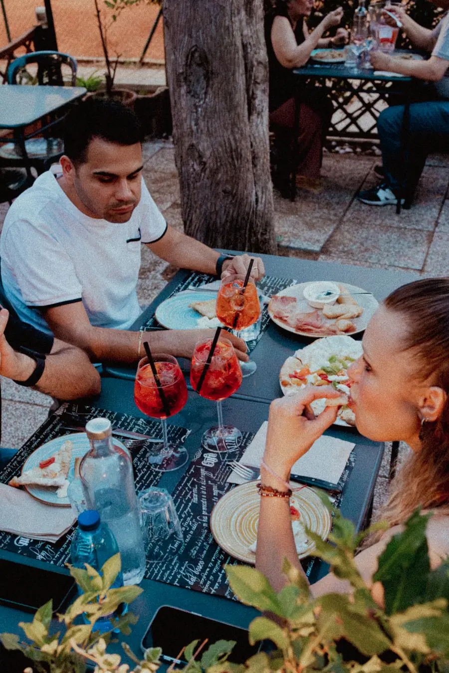 Due clienti a cena a un tavolo del dehors, con altri tavoli occupati e gli alberi alle spalle