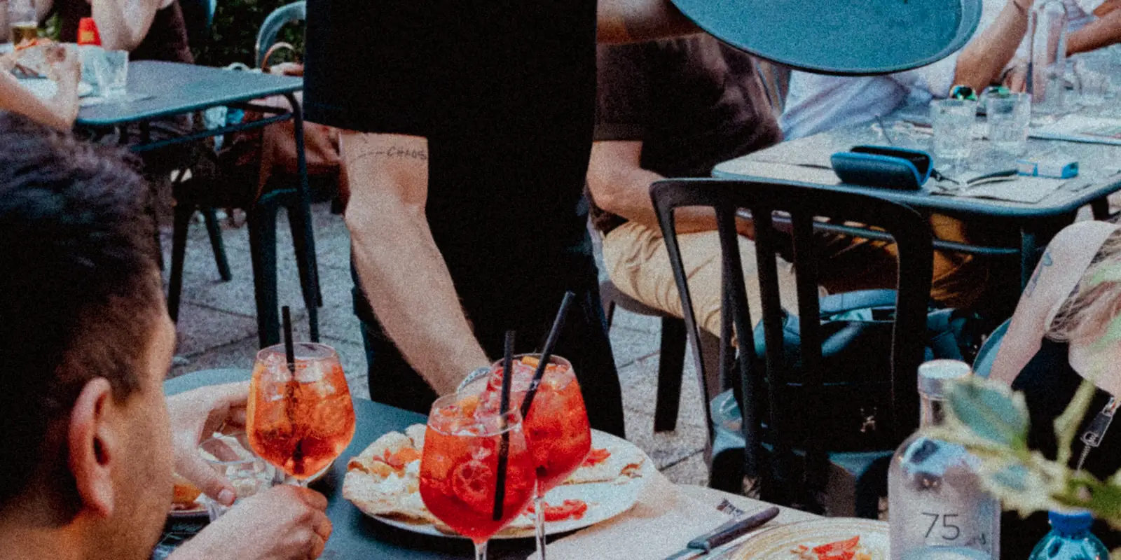 Servizio ai tavoli del dehors: spritz e piatti su un tavolo, altri clienti seduti sullo sfondo